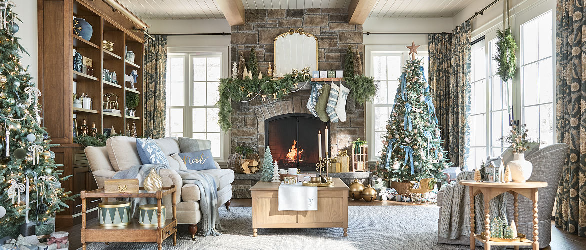 Un salon lumineux et festif décoré pour Noël, comprenant un sapin de Noël orné de décorations à côté d’un foyer allumé. Des guirlandes de Noël, des ornements, des décorations de table et des accessoires saisonniers créent un décor des Fêtes partout dans l’espace.