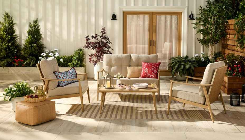 A neutral toned patio set with a love seat and two chairs on a backyard patio surrounded by plants.