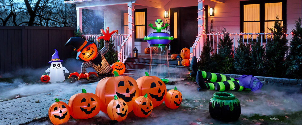 A front yard Halloween display featuring illuminated inflatable jack-o’-lanterns, a giant pumpkin-headed character, a ghost with a witch hat holding a trick-or-treat bucket, a floating alien decoration, a witch’s striped legs beside a cauldron in front of a house decorated with orange string lights.
