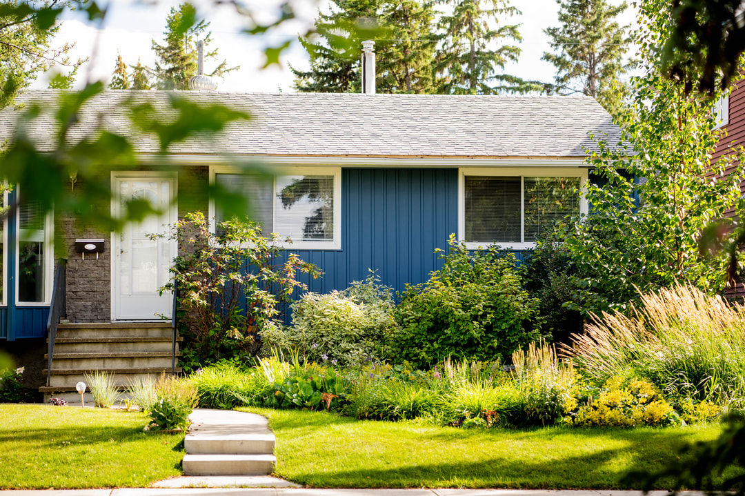 A beautifully maintained blue bungalow featuring mature landscaping, vibrant greenery, and a welcoming front entrance. This residential home exterior showcases a peaceful yard with shrubs, ornamental grasses, and a sunny, well‑kept lawn.