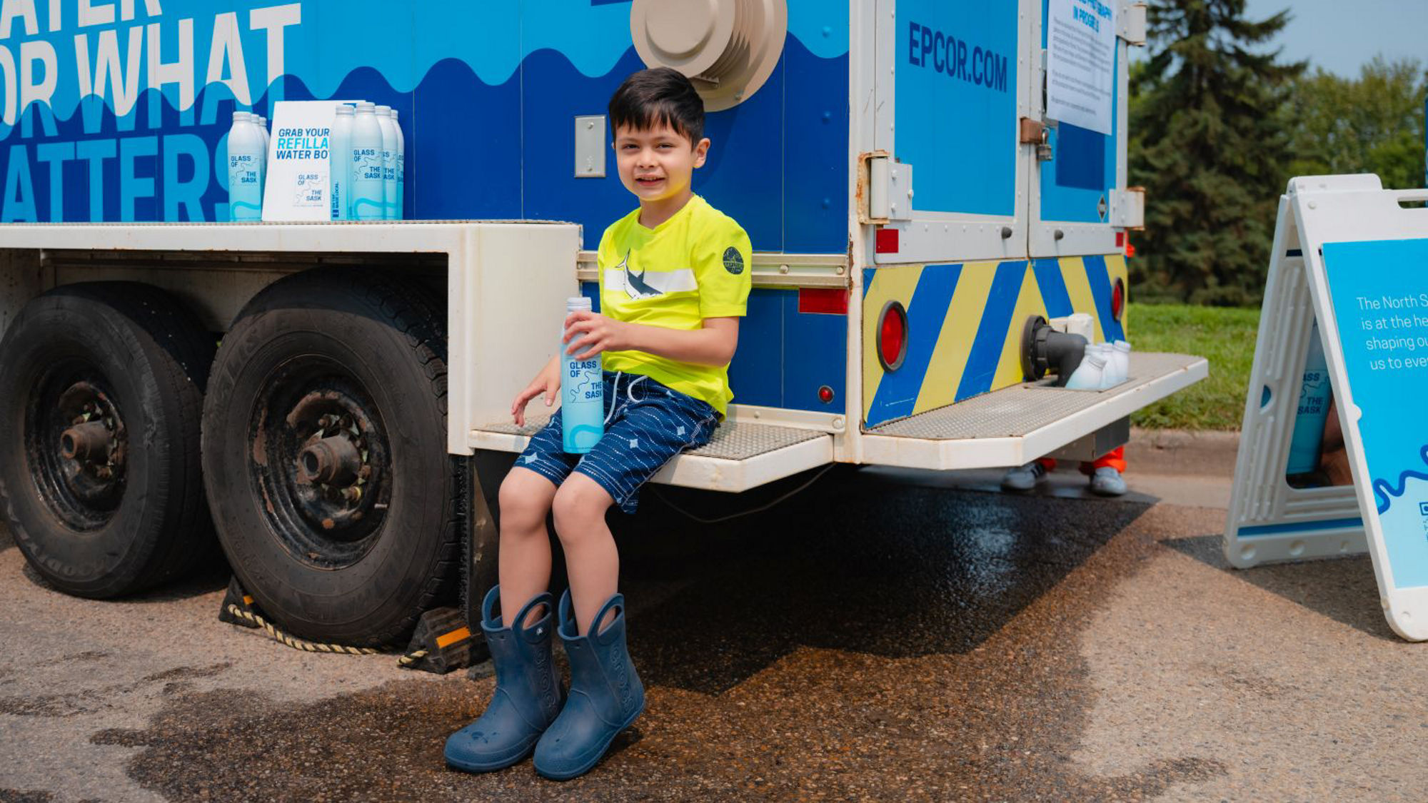 A young child smiling while sitting on an EPCOR water tank after filling up a Glass of the Sask aluminum bottle.