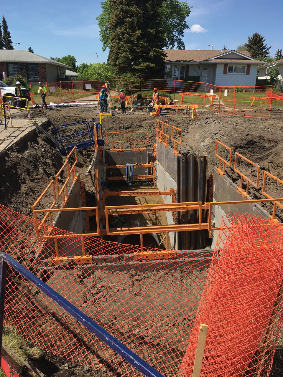 Utility crew wearing safety gear working on an excavated trench with steel and wood shoring, with residential homes located in the background.