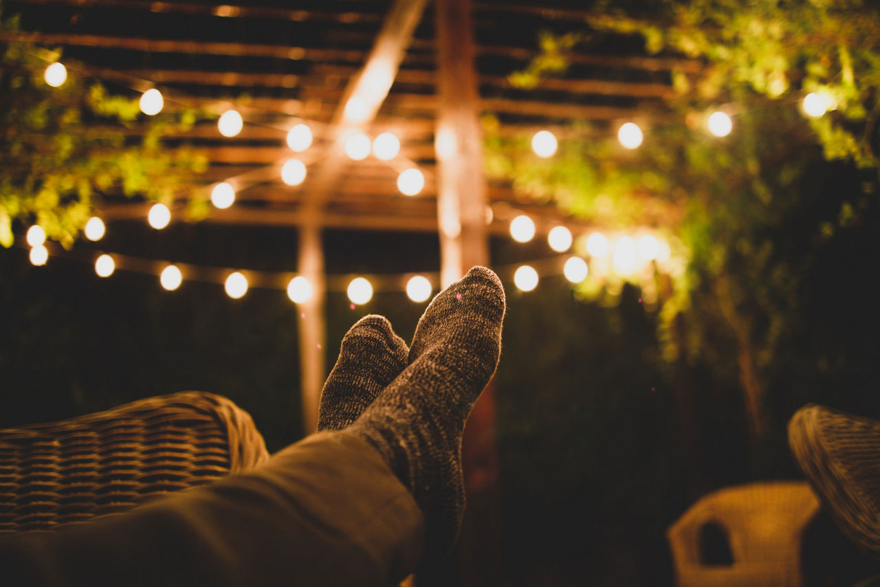 A relaxed nighttime scene showing a person resting with socked feet in the foreground, surrounded by warm glowing string lights and greenery, creating a cozy backyard or patio atmosphere.