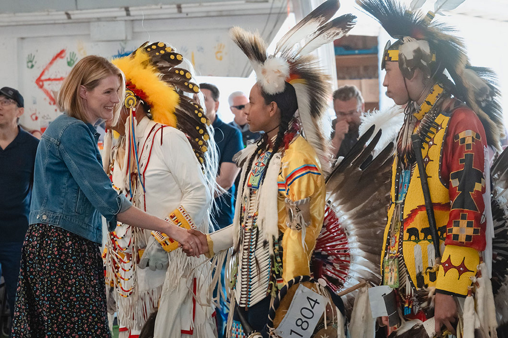 Participants greet one another during a community pow wow, with individuals wearing detailed traditional regalia inside a gathering space that celebrates culture, ceremony, and connection.