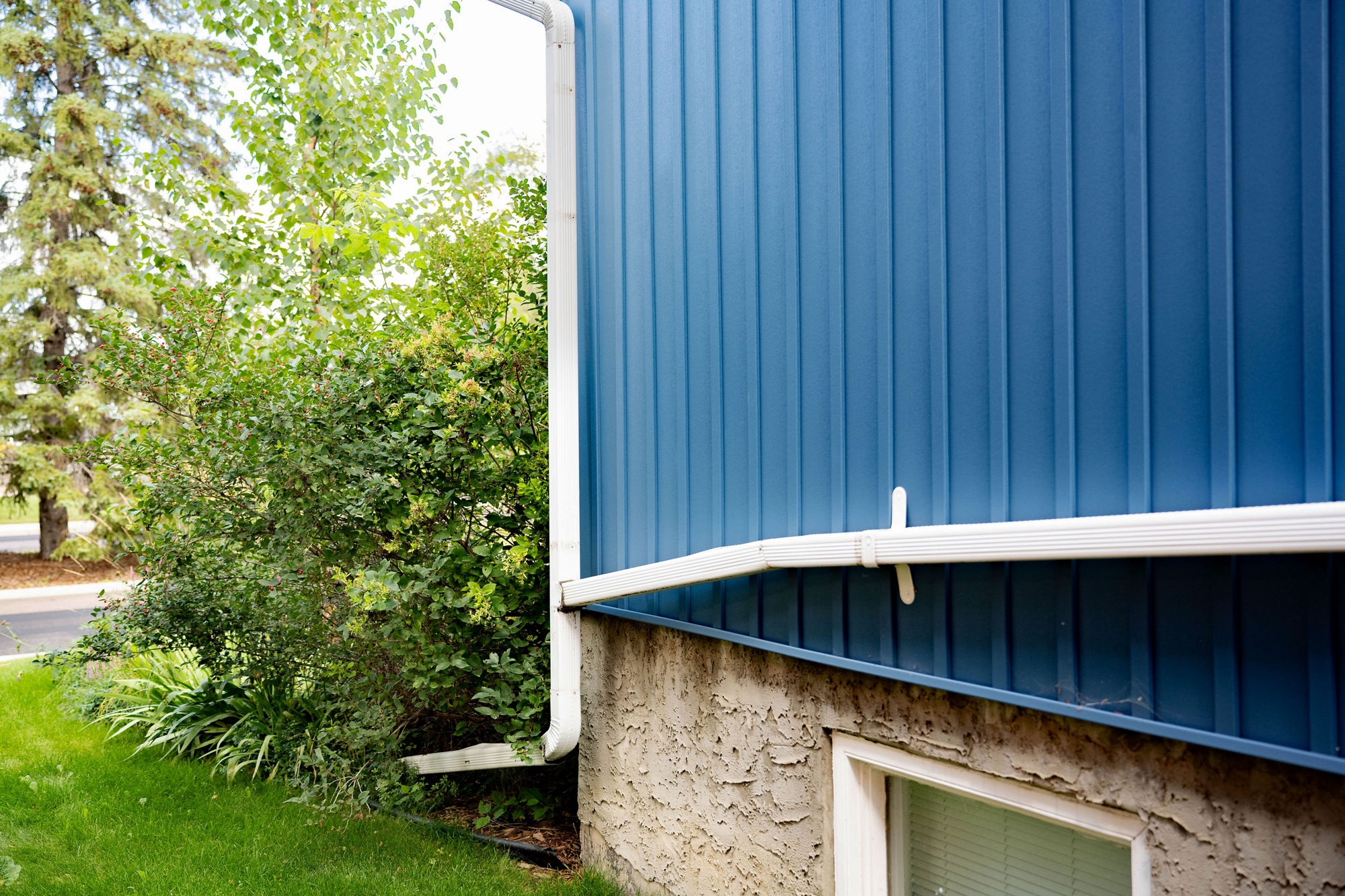 A long white downspout running horizontally along the side of a blue‑sided house before turning downward toward the ground. The downspout leads runoff away from the building’s foundation, with shrubs, trees, and lawn visible around the structure.