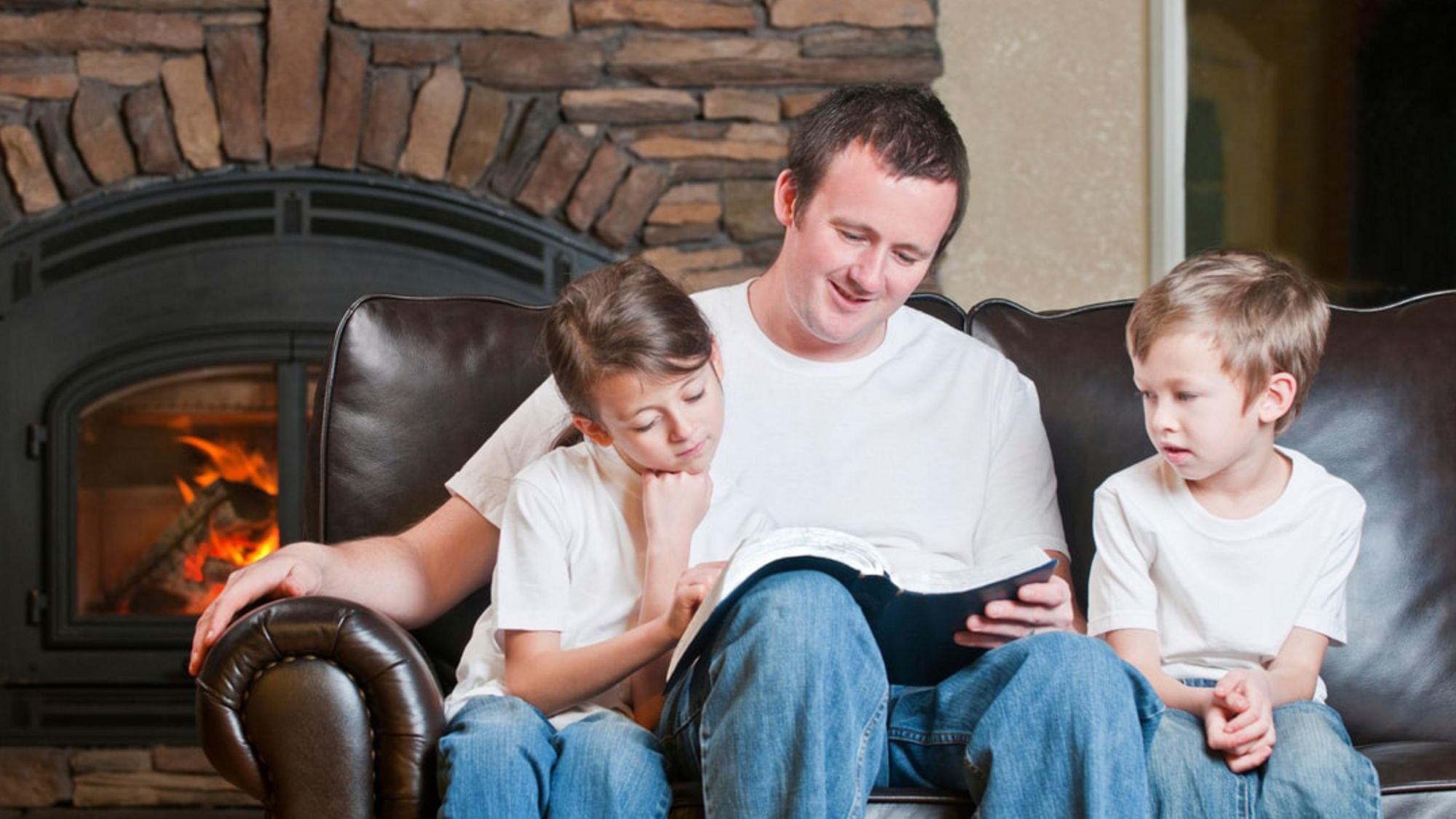 A father sits on a couch reading a book with his two children beside a lit fireplace, showing a calm family moment in a cozy home setting.