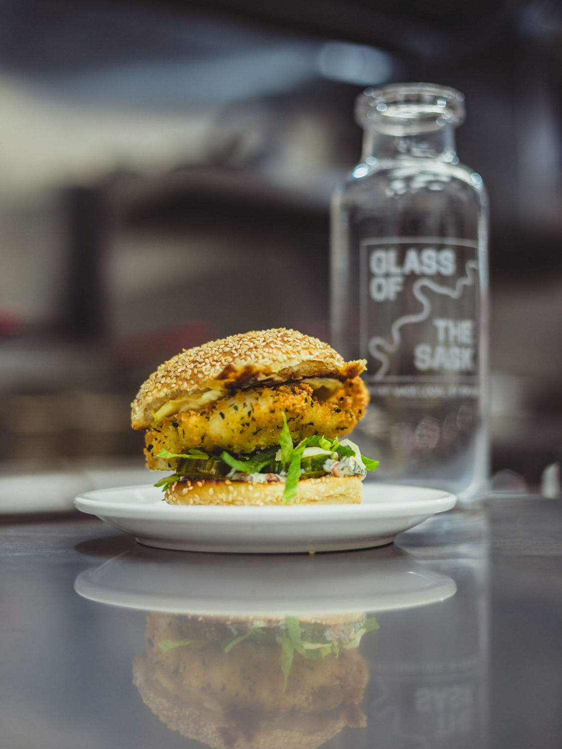 Crispy fried Atlantic cod sandwich with fresh lettuce and pickles on a sesame seed bun, plated on a white dish with a branded Glass of the Sask carafe in the background.