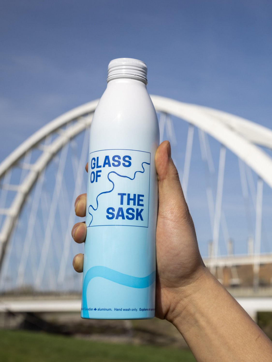 Someone holding a Glass of the Sask bottle with the river behind it, and an Edmonton bridge in the distance