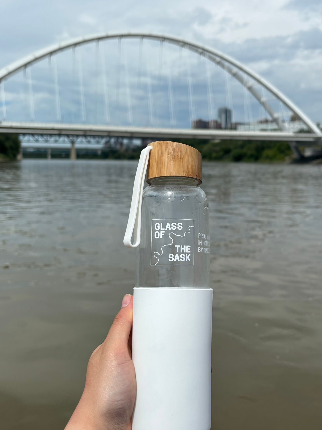 Someone holding a Glass of the Sask bottle with the river behind it, and an Edmonton bridge in the distance