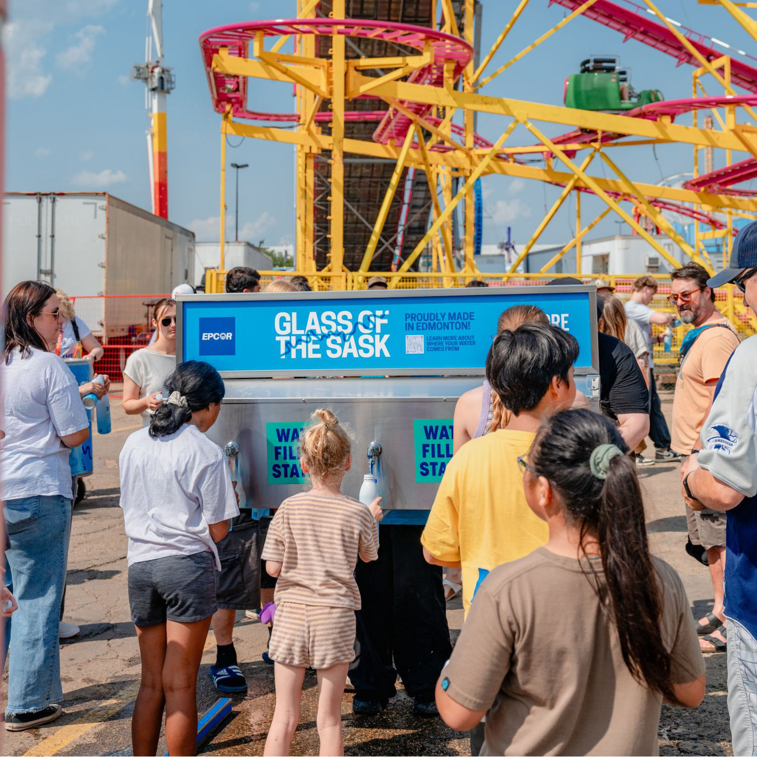 Children lined up at a Glass of the Sask water fill station near a pink and yellow rollercoaster at KDays.