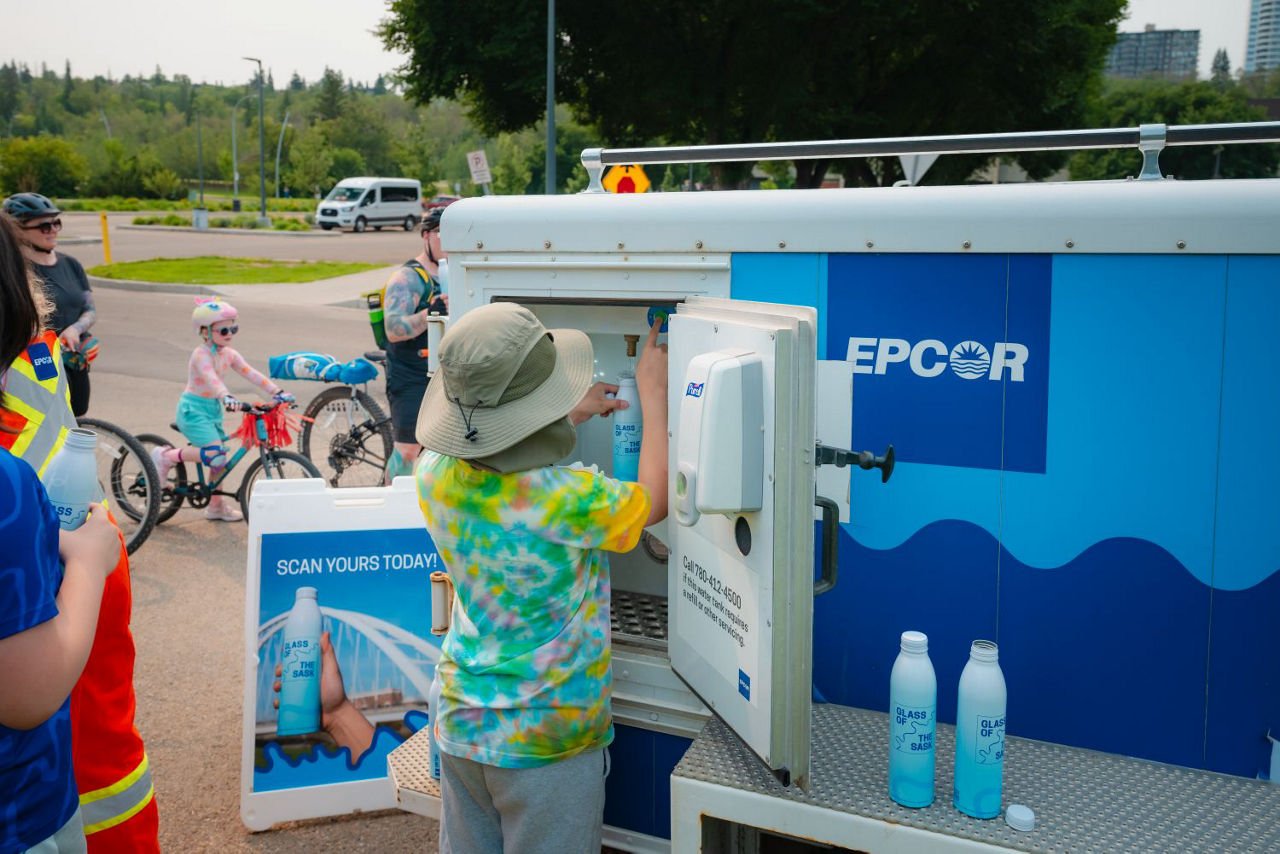 A child in a tie dye shirt and hat filling up aluminum Glass of the Sask bottle during an outdoor event at an EPCOR water tank.