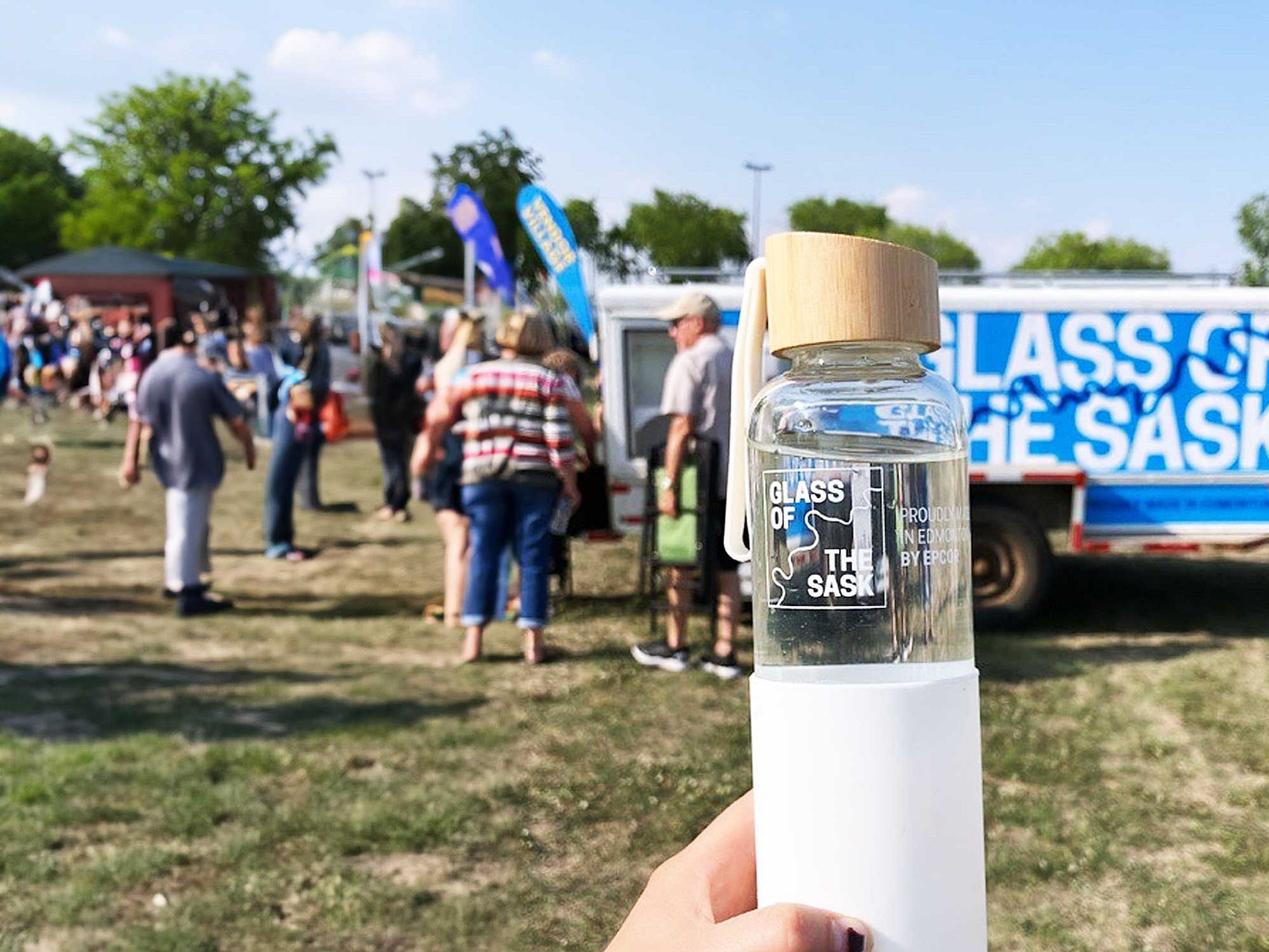 A Glass of the Sask water bottle being held up in front of the water tank at a festival.