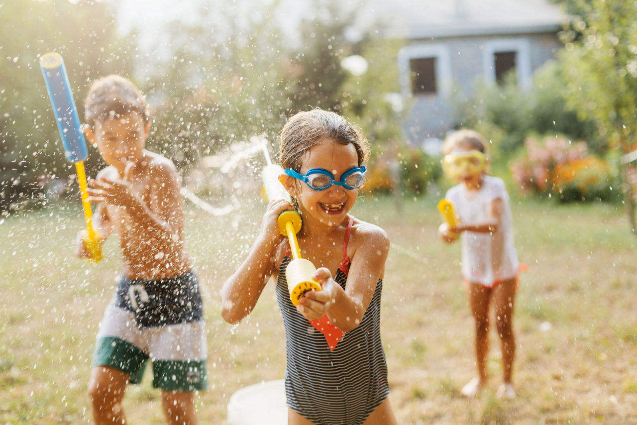 Children play outdoors on a sunny day, splashing water and using colorful water guns in a backyard setting.