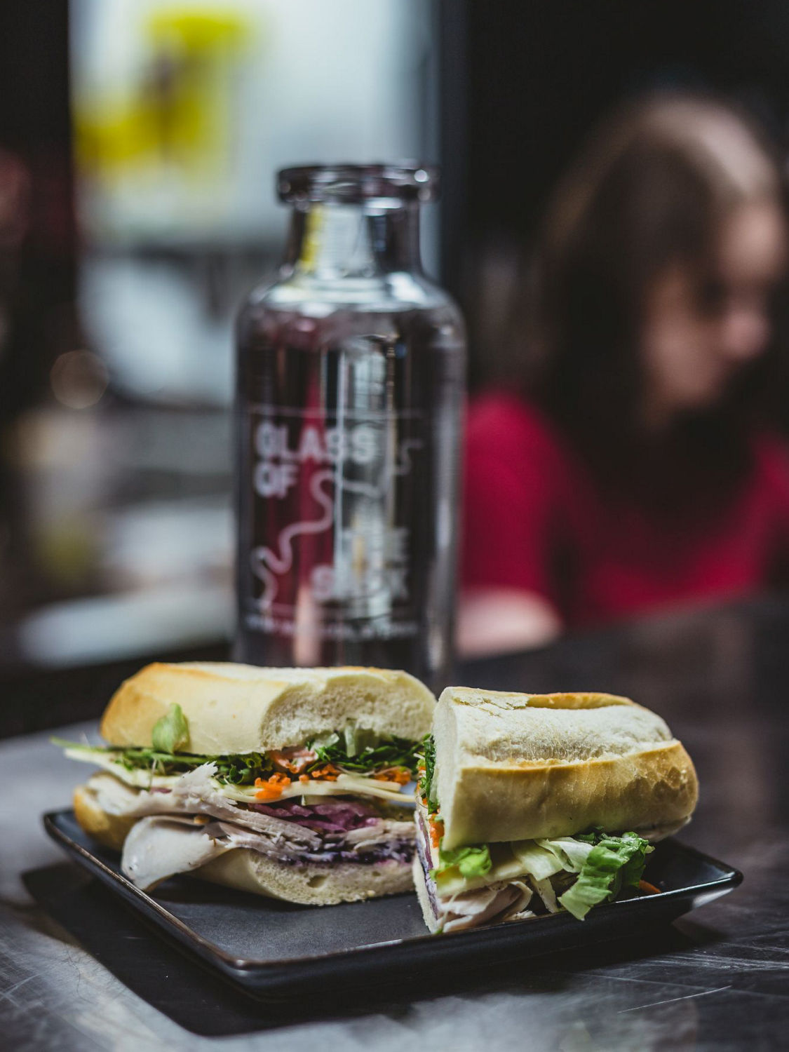 Fresh house-roasted turkey sandwich served on a panini-style baguette, sliced and presented on a black plate, with a Glass of the Sask carafe displayed in the background.