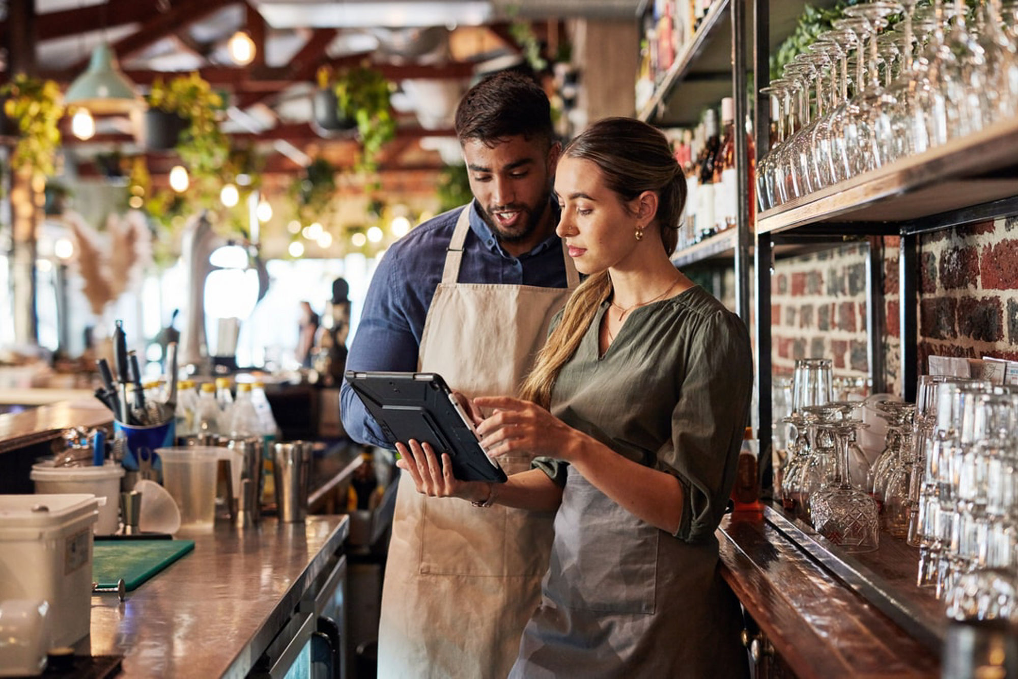 Two people stand behind a café or bar counter reviewing information on a tablet, with glassware and shelves visible in a warmly lit interior.