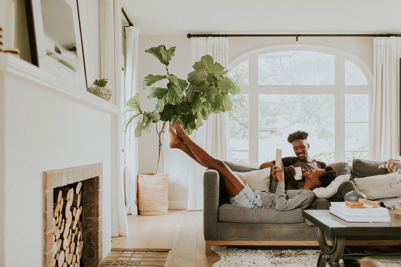 Two people unwind on a comfortable sofa in a bright, modern living room featuring large windows, natural light, and indoor plants.