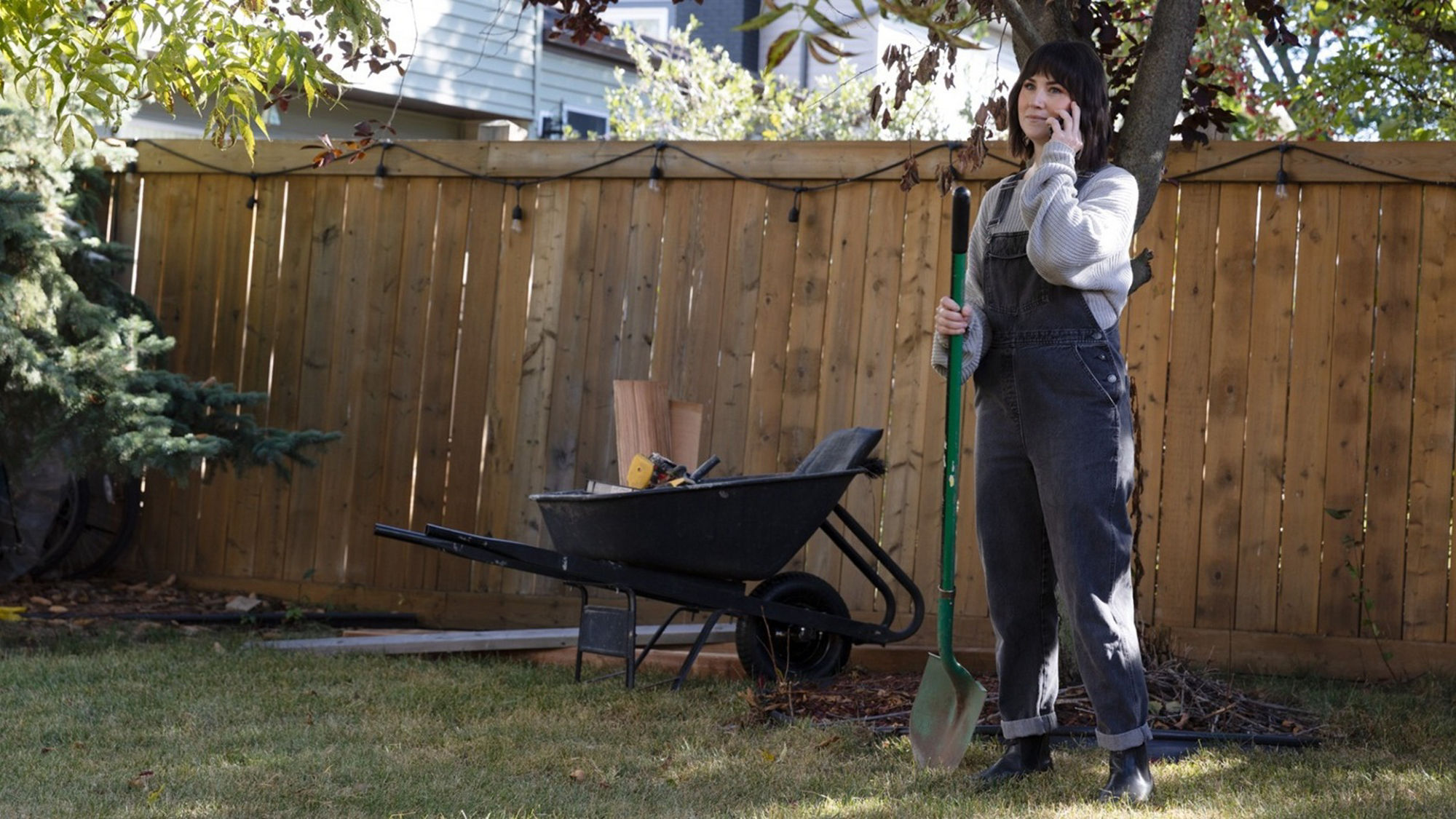 Woman standing in a backyard holding a shovel and talking on the phone beside a wheelbarrow filled with tools near a wooden fence.