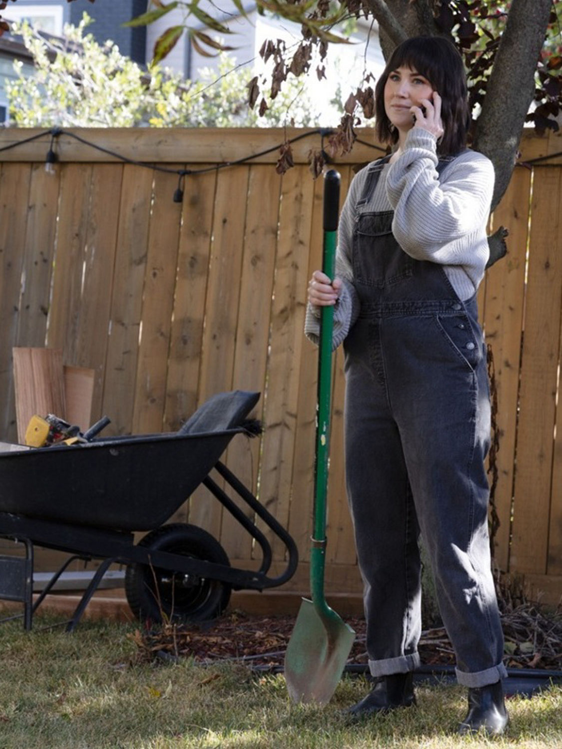 Woman standing in a backyard holding a shovel and talking on the phone beside a wheelbarrow filled with tools near a wooden fence.