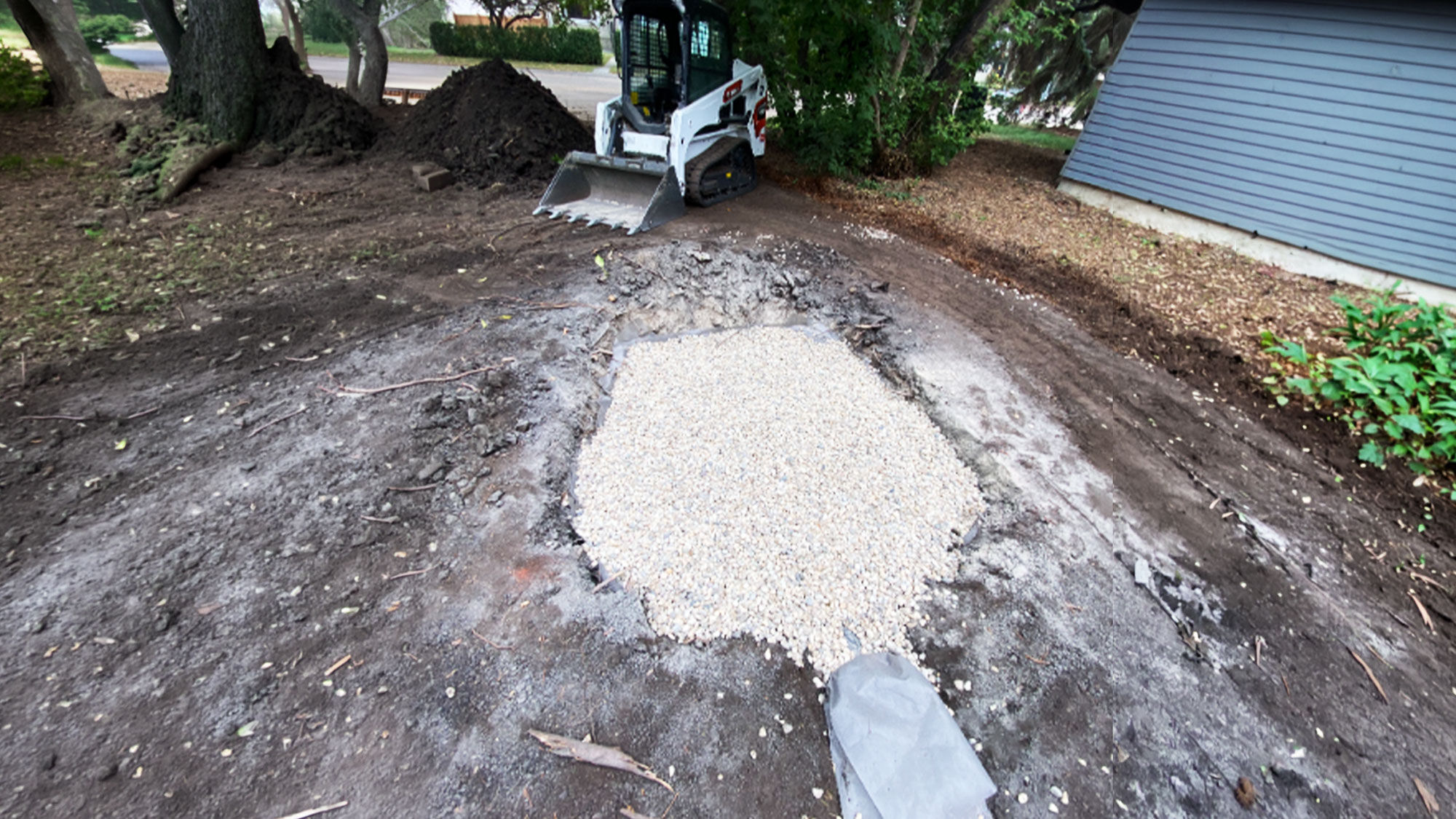 A soakaway pit is excavated and partially filled with gravel, with a compact track loader positioned nearby in a residential yard during landscaping or drainage work.