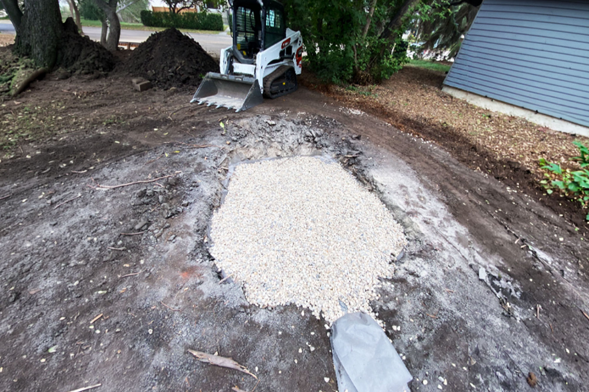 A soakaway pit is excavated and partially filled with gravel, with a compact track loader positioned nearby in a residential yard during landscaping or drainage work.