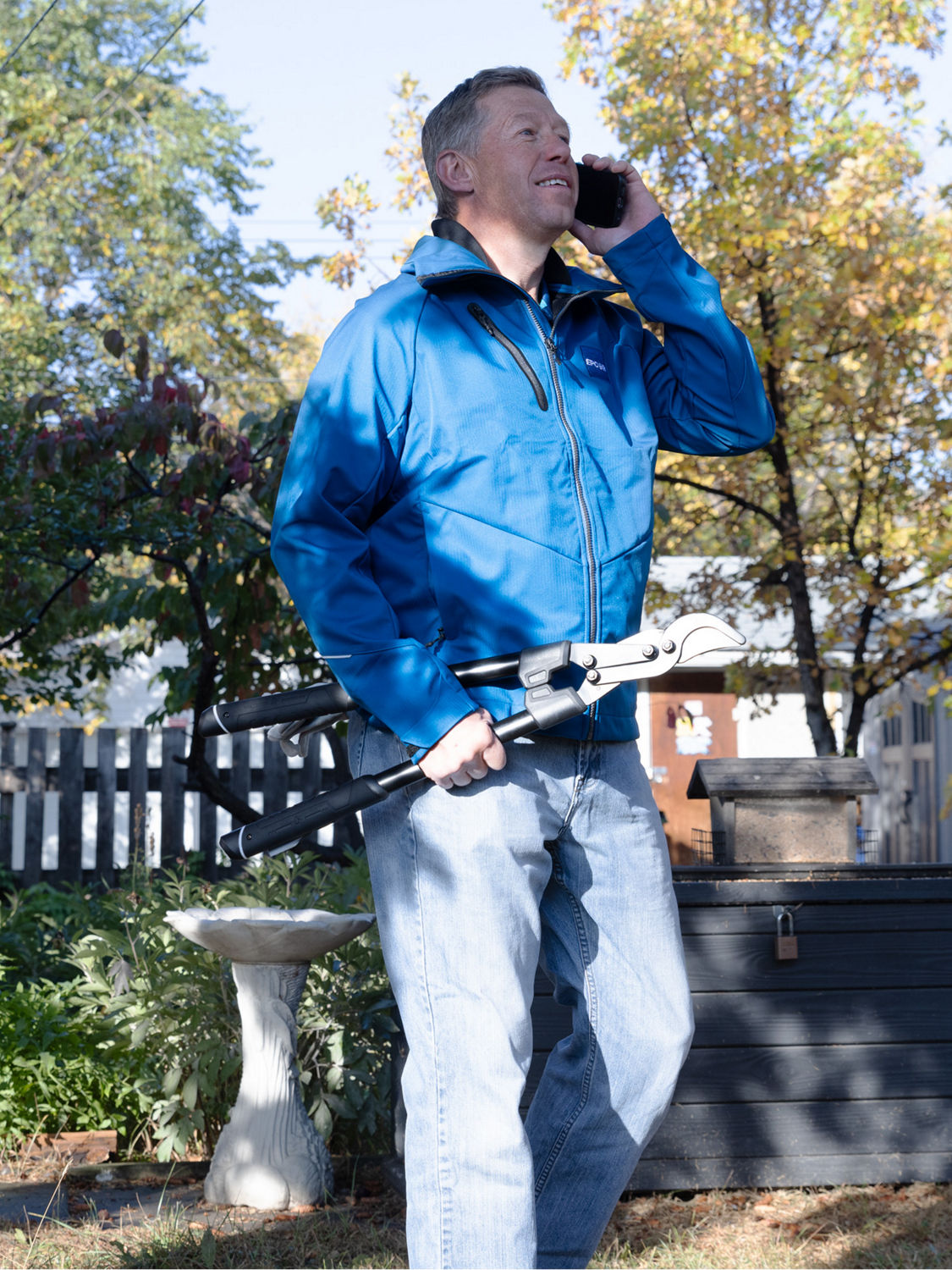 A person holding tree trimmers in one hand is talking on the phone in their yard.