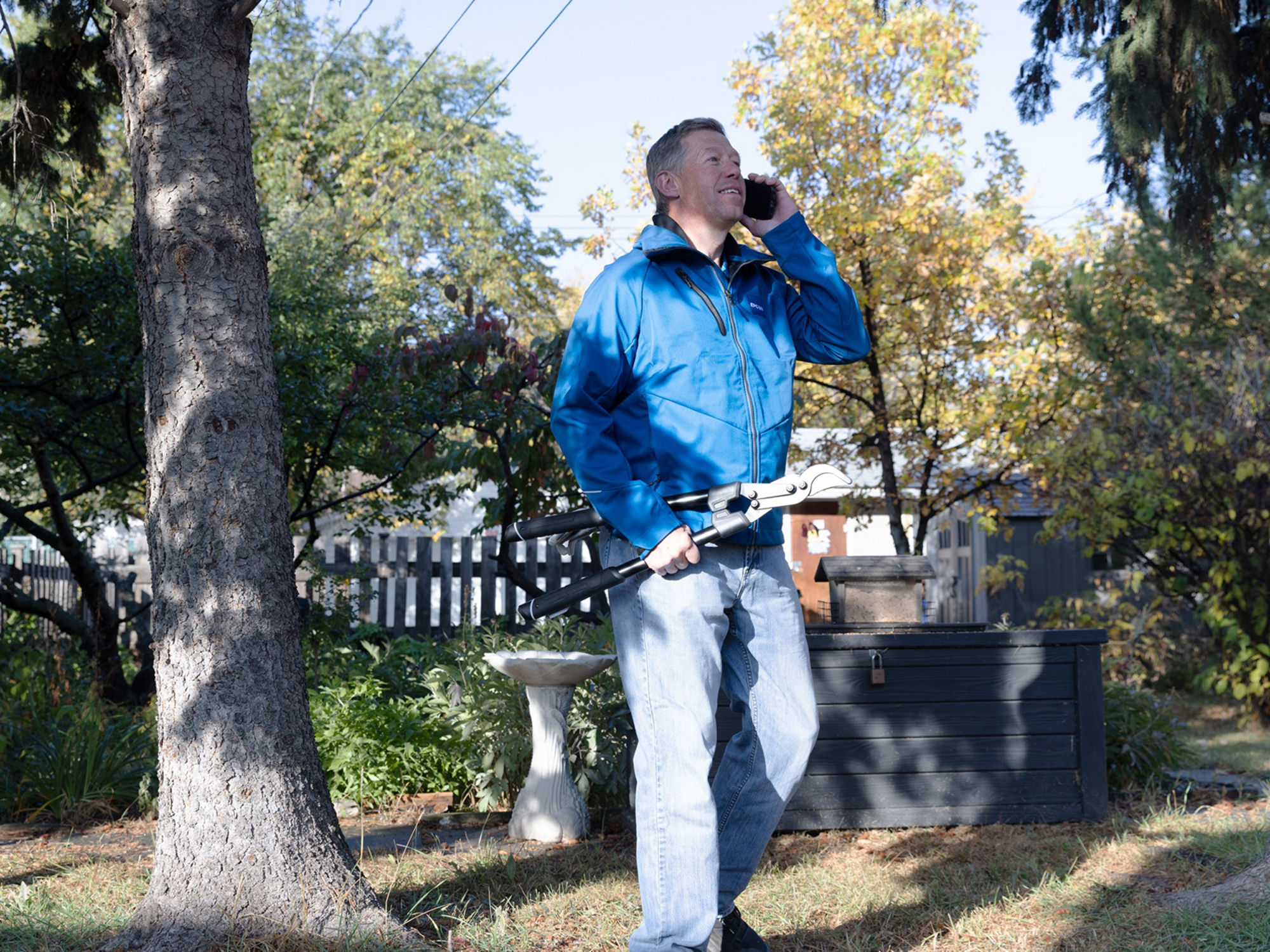 A person standing in their yard holds pruning shears while speaking on a phone.