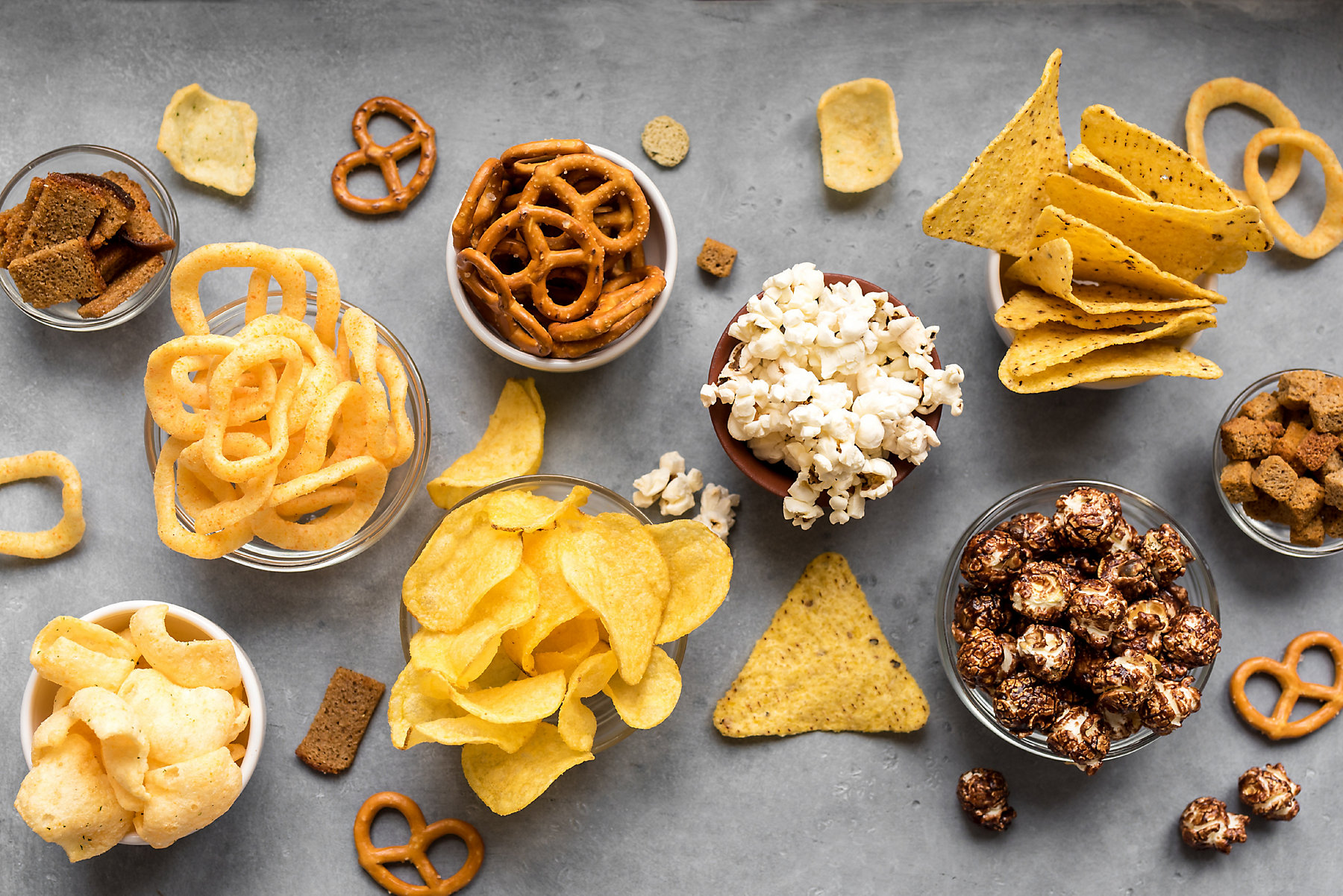 Assortment of Unhealthy Snacks: chips, popcorn, nachos, pretzels, onion rings in bowls, top view