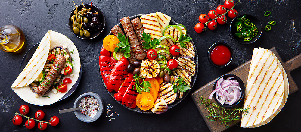 Grilled meat kebabs, vegetables on a black plate with tortillas, flat bread. Slate background. Top view.