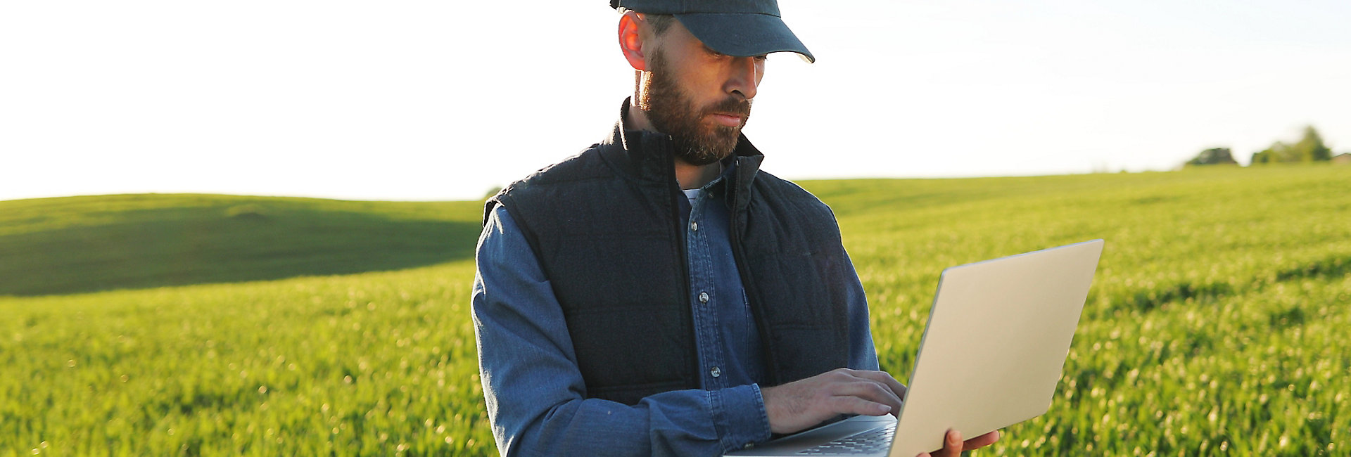 Caucasian young male farmer in hat standing in field and typing on keyboard of laptop computer. 