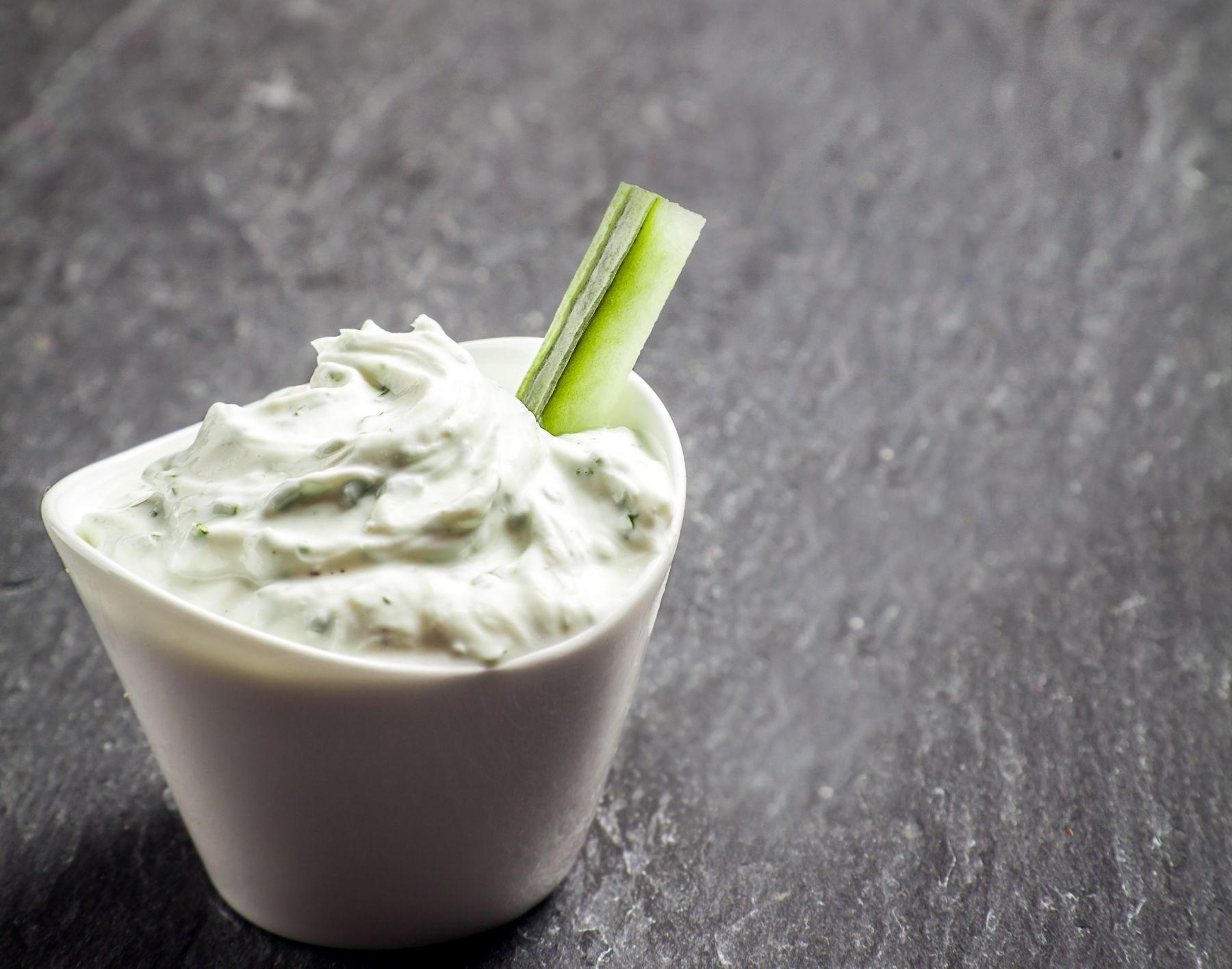 Delicious creamy vinaigrette and cottage cheese sauce in a ceramic dish with a cucumber crudites or baton for a healthy snack or appetizer on grey slate with copyspace