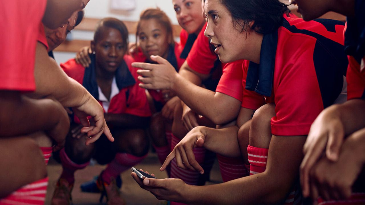 A female football team huddled around a phone in the locker room.
