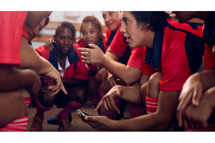 A women’s football team huddling in the locker room, game planning with a phone in one player’s hand. 