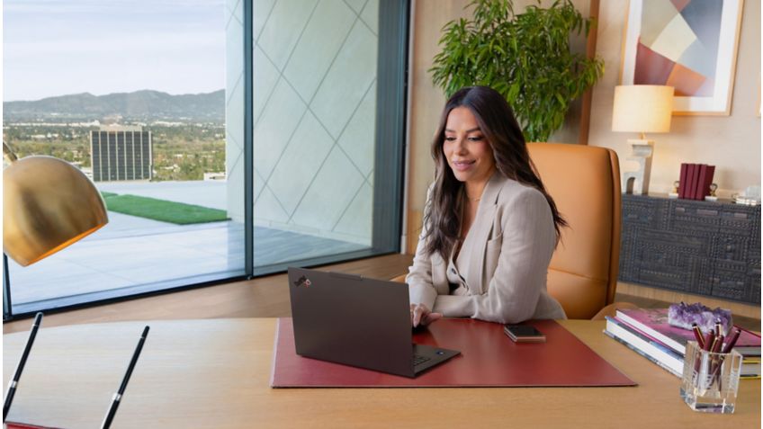 エヴァ・ロンゴリアはデスクの後ろに座り、パワースーツを着ている。彼女は前にある開いたThinkPad X9 15p G1をコミカルに指さす。スクリーンセーバーは、赤いカーペットの上で華やかな赤いドレスを着た彼女自身の画像です。 