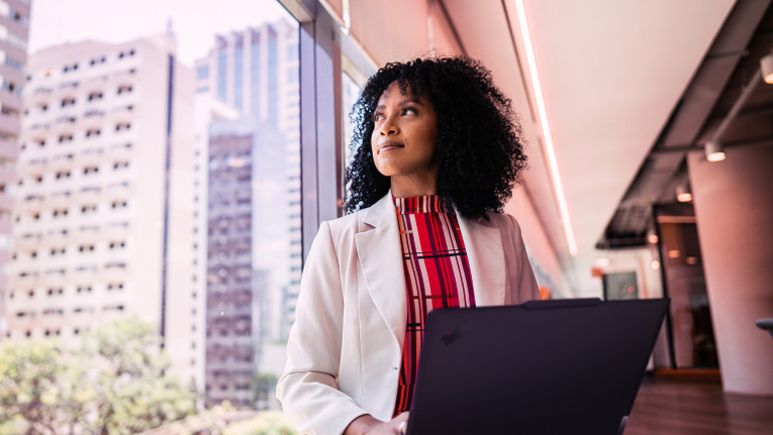 Une femme utilise son portable à côté d’une fenêtre, un gratte-ciel est derrière elle au premier plan.