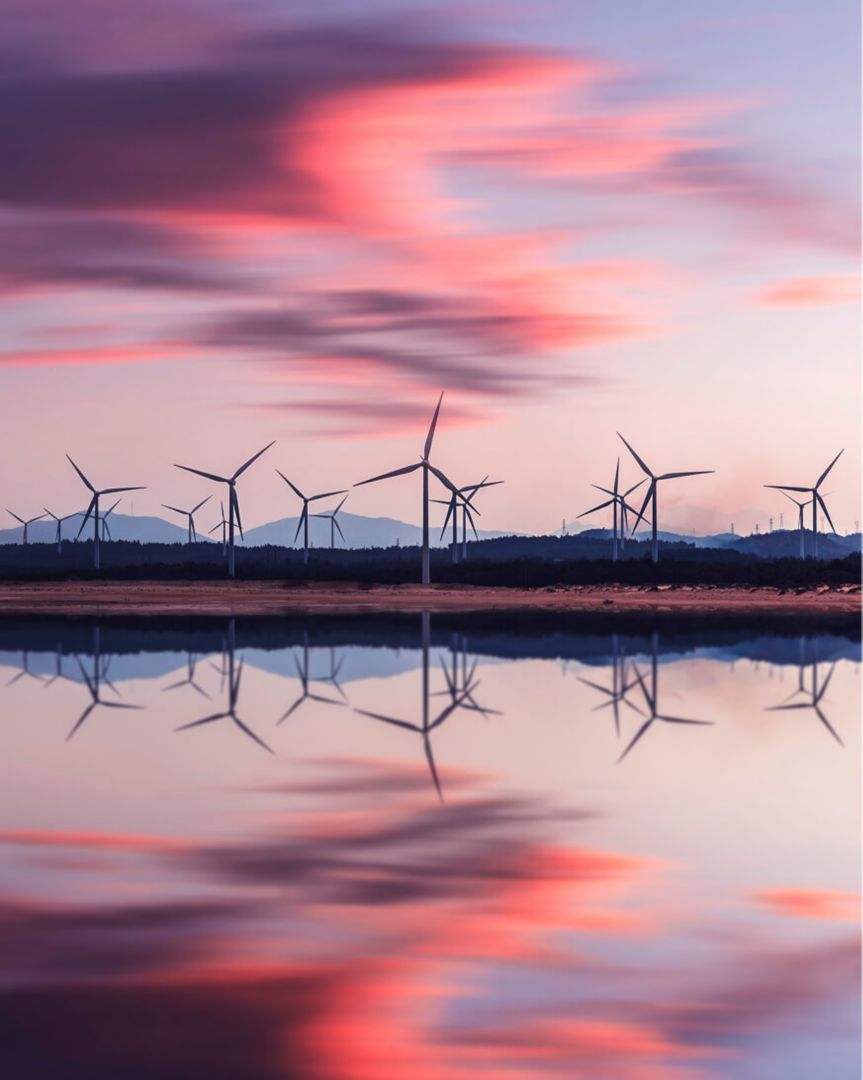 A lake with wind turbines and mountains in the background under a rosy-colored sky at dusk.