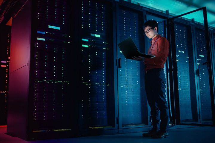 A man standing in a server room and holding a laptop. 