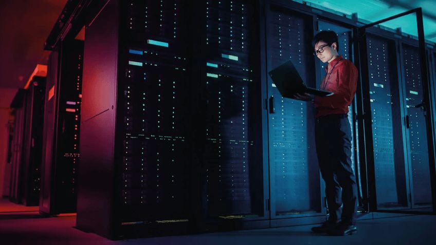 Technician with a laptop in a server room lit by red and blue lights.
