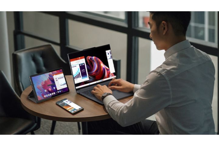 A man in a white dress shirt sits at a round wooden table, working on a Lenovo laptop, tablet, and Motorola smartphone. All three devices display active business applications and colorful interfaces.