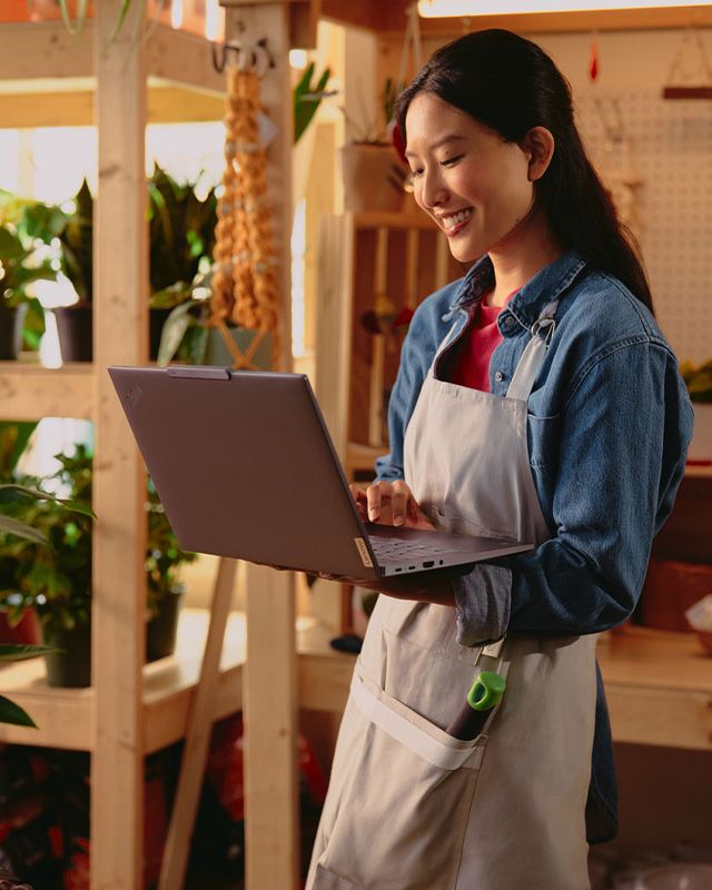 Woman stands in a plant shop typing on a Lenovo ThinkPad with one hand.