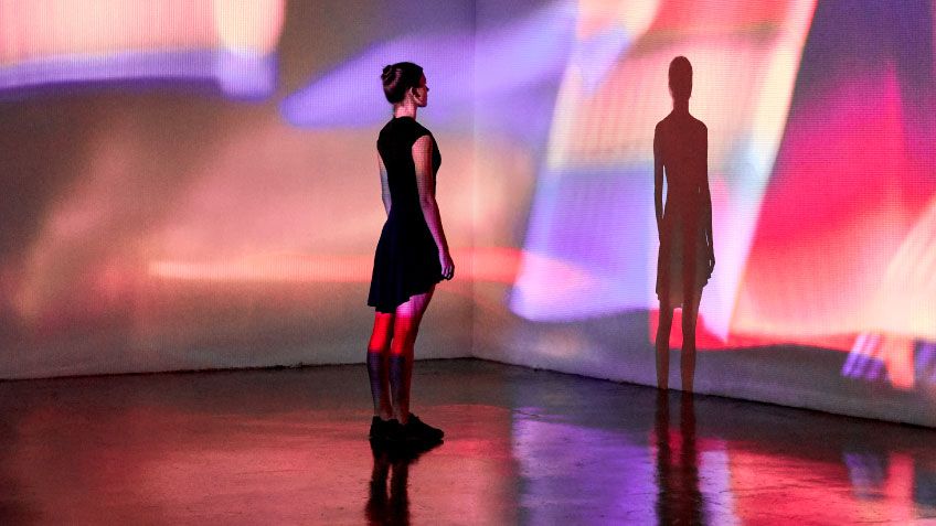 Person in a black dress stands in a colourful digital art installation with vibrant projections on walls and a reflective floor.