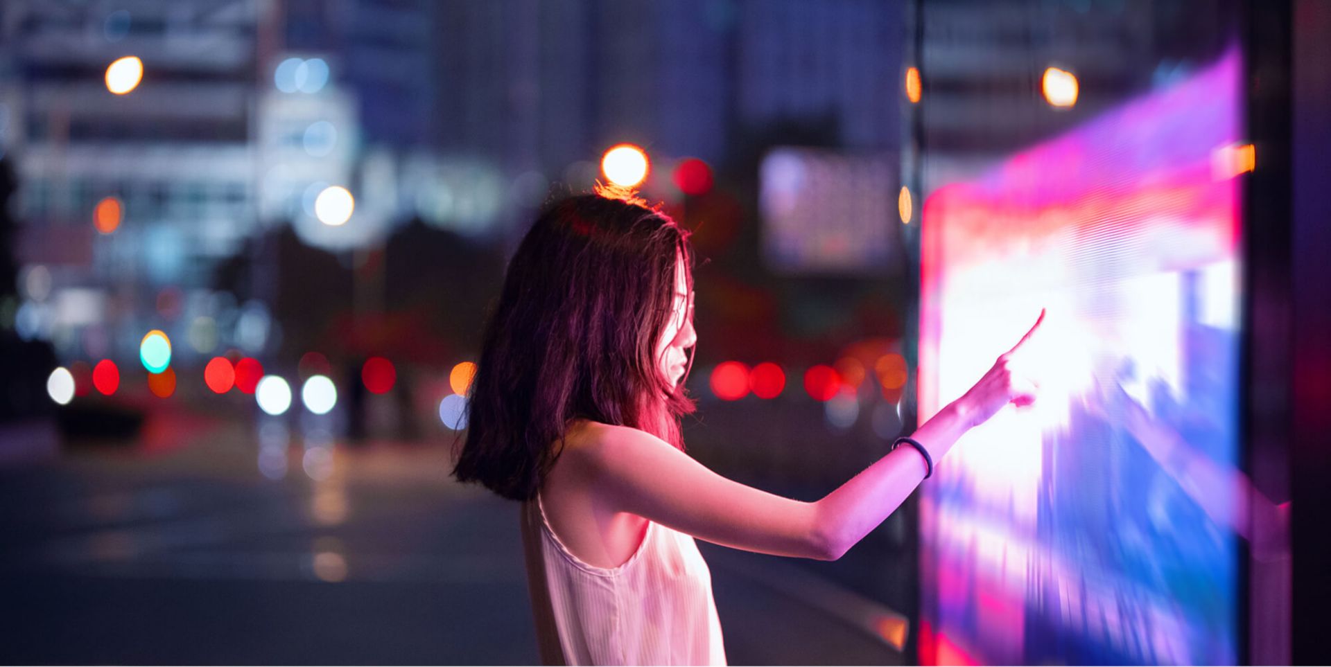 A girl holding her finger up to a touch screen in a city at night.