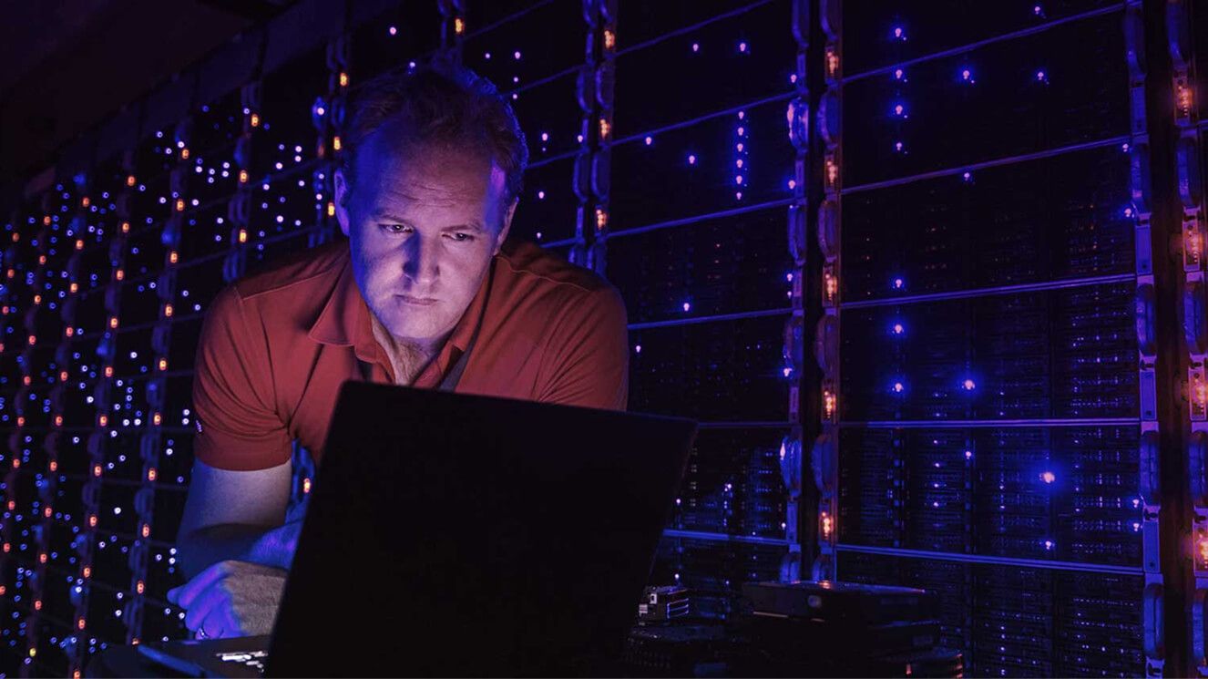 A man looking at his computer in a dark server room. 