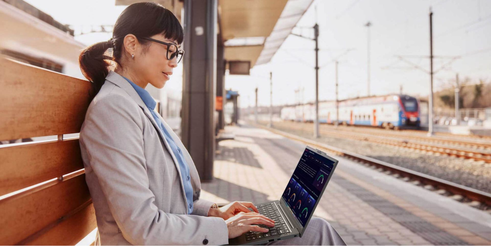 A female using her laptop while seated on a bench at an outdoor train station. 