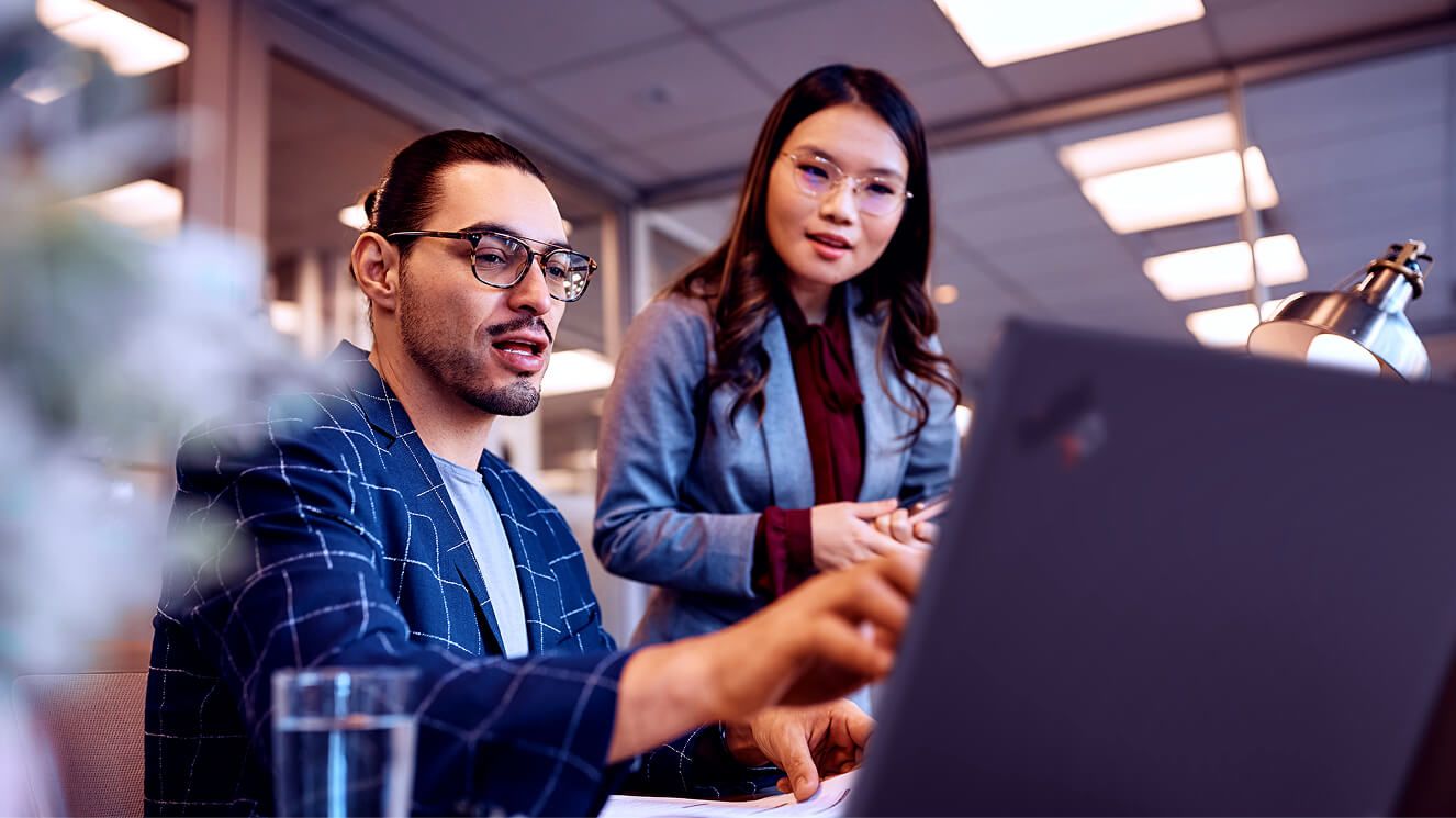 A colleague assisting her male coworker who is working on his laptop. 