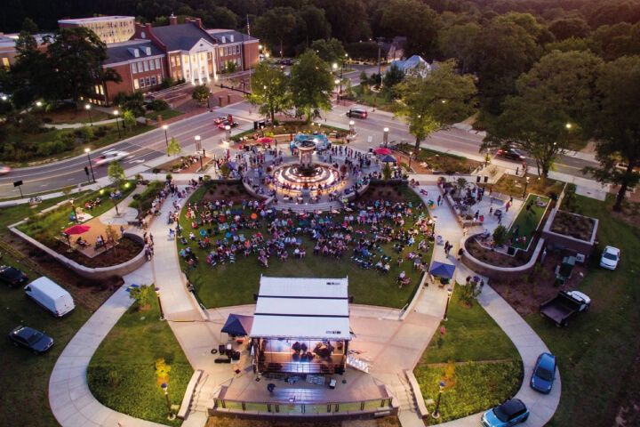A crowded park with a fountain and stage during an evening event.