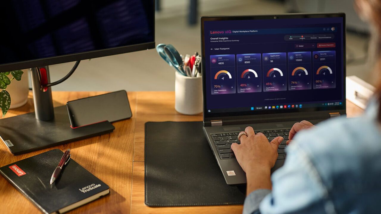 Una mujer escribiendo en una laptop en su escritorio, con una segunda pantalla y un cuaderno a su lado.