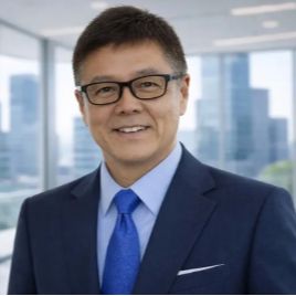 黒い長方形の眼鏡をかけ、紺色のスーツ、淡い青色のシャツ、鮮やかな青色のネクタイを着用した笑顔の男性が、大きな窓のあるモダンなオフィスに立っている。背景には街のスカイラインが見える。