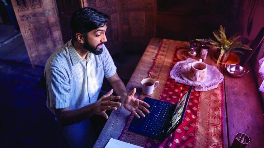 A bearded, dark-haired person holds a cup of coffee as he talks during a video call on his laptop, which sits on a wood table with red and white fabric coverings.