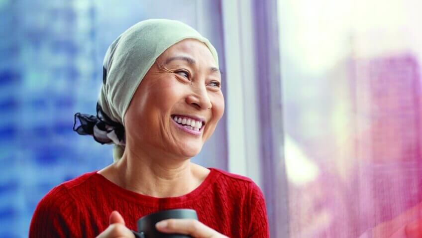 赤いシャツを着て頭に淡い薄緑の布を巻いた女性が窓から彼女の左手にある都会の高層ビルを見ている。