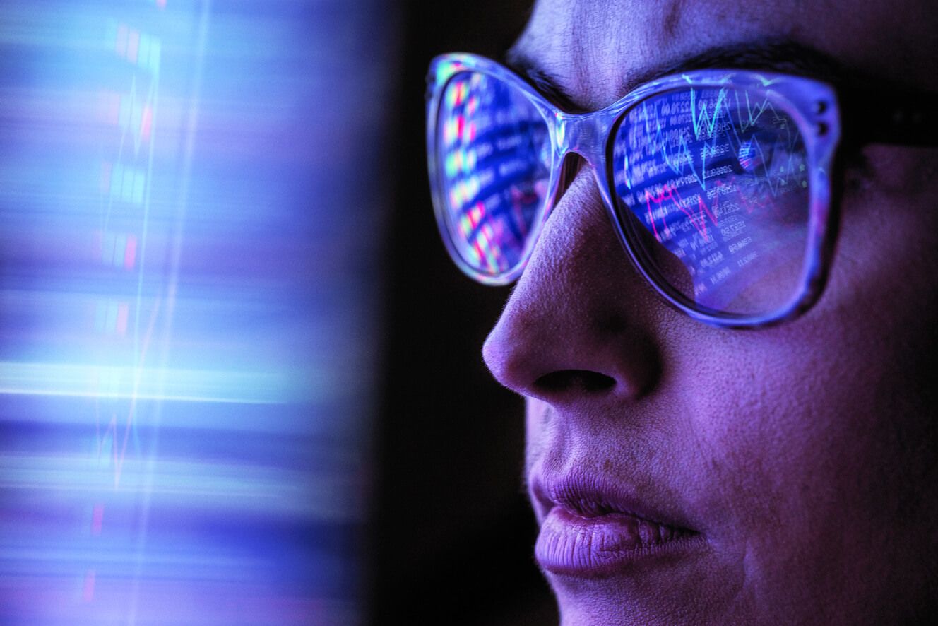 A woman looking at a computer screen and the data reflecting in her glasses.  